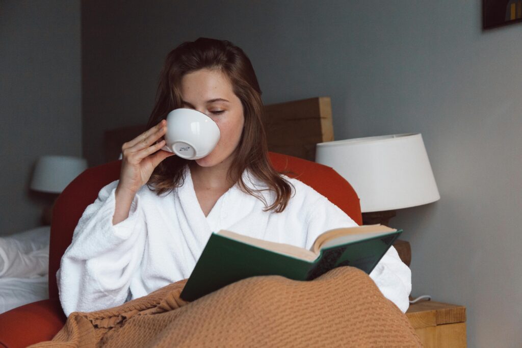 Image of woman in bed drinking a cup of tea and reading a book to prevent burnout